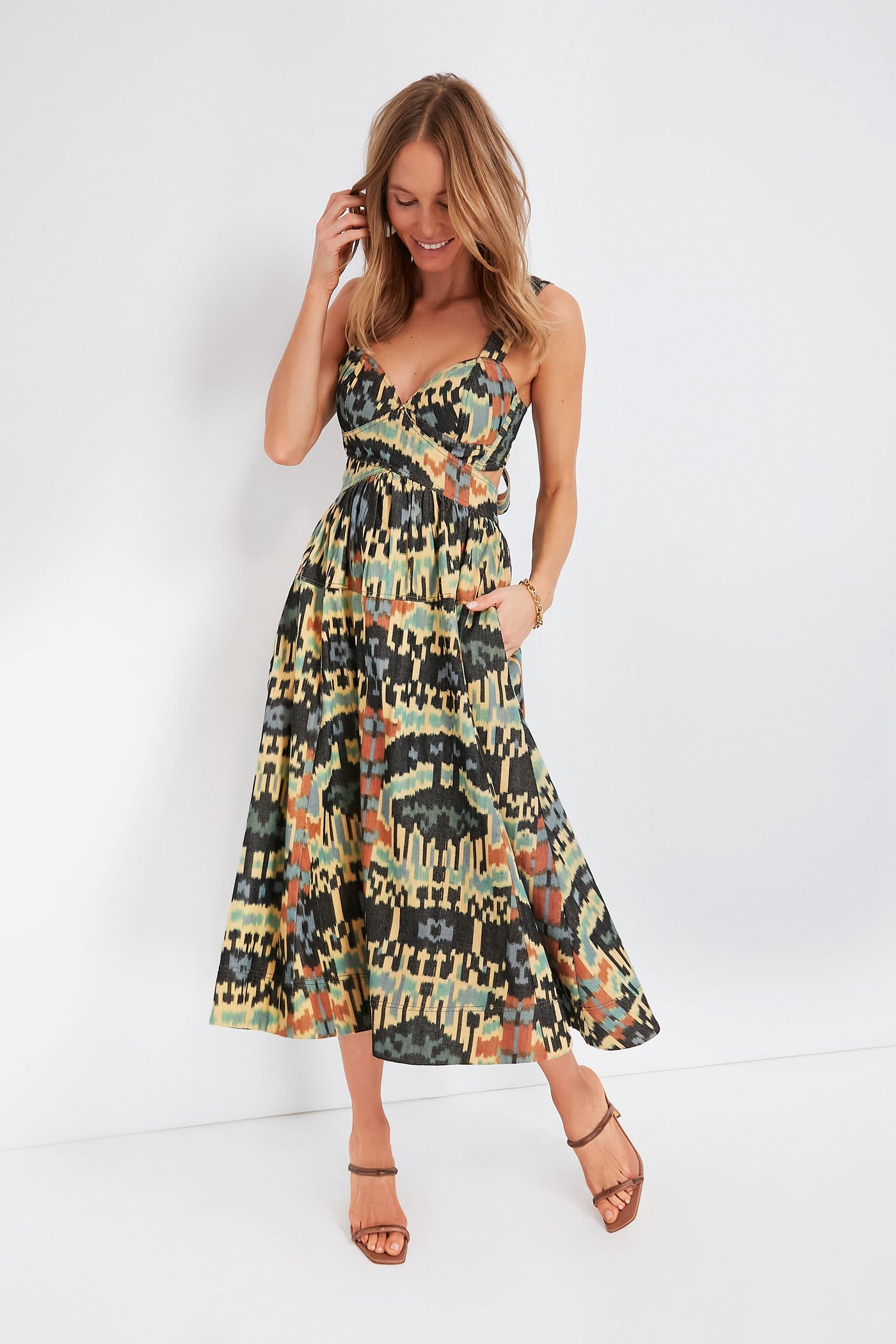 A woman smiles and touches her hair while wearing a sleeveless Ulla Johnson dress with thin straps, paired with strappy sandals.