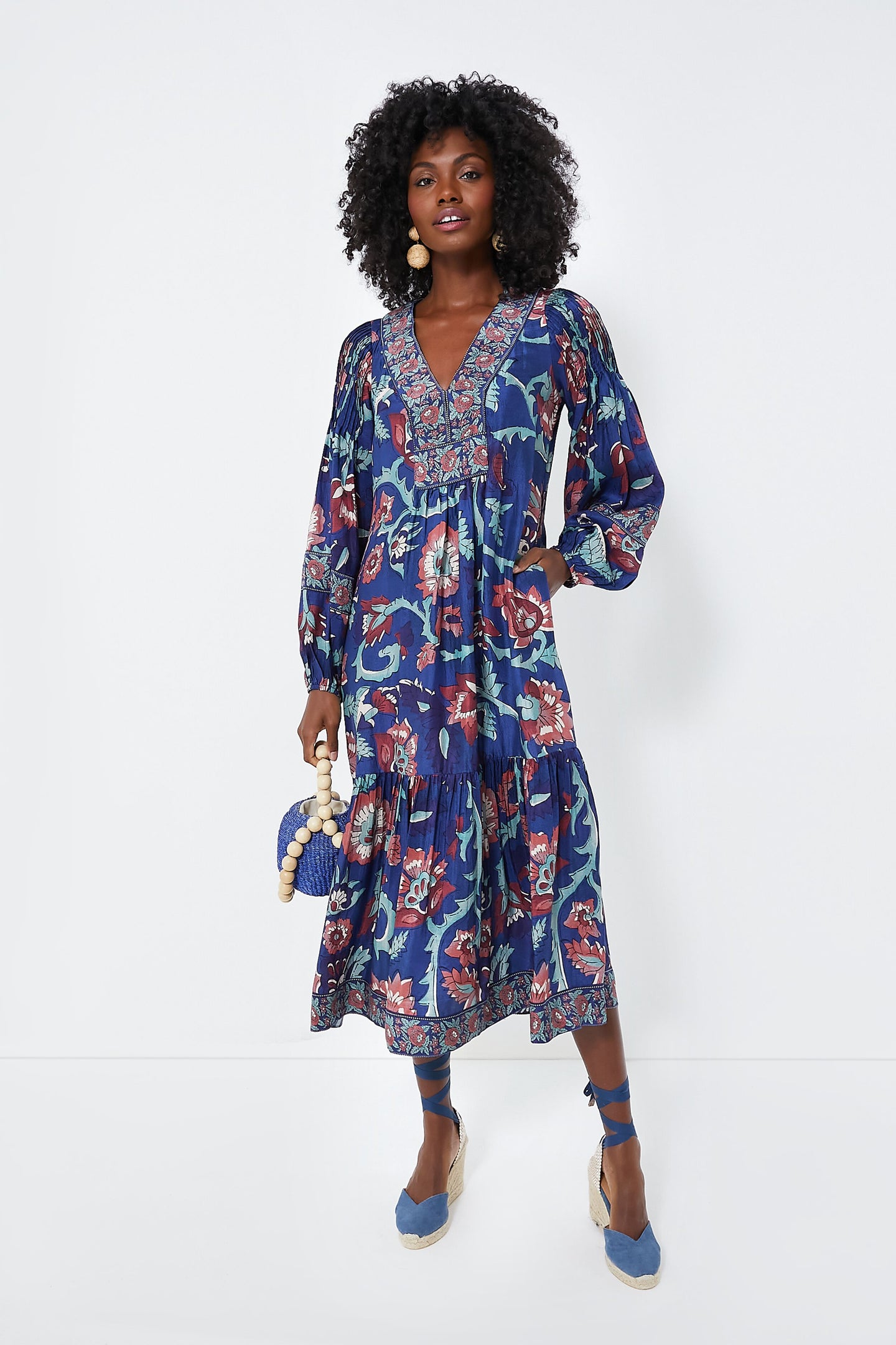 A woman in a Sea New York Rory Print Dress with long sleeves and a V-neckline, carrying a small blue bag and wearing blue ankle-tie espadrilles.