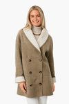 A woman wears a Tuckernuck tan double-breasted suede coat with white shearling collar and cuffs over a cream turtleneck sweater and necklace, smiling. A chic winter outerwear choice.