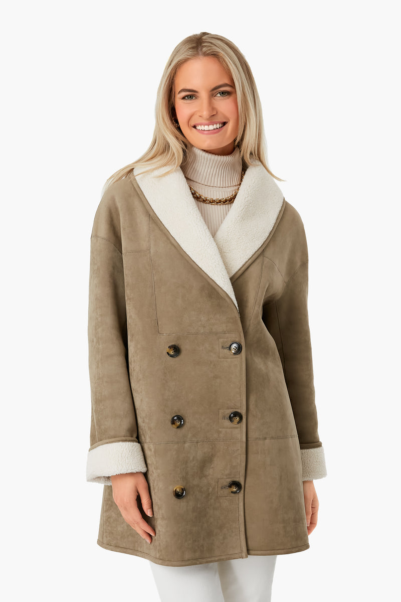A woman wears a Tuckernuck tan double-breasted suede coat with white shearling collar and cuffs over a cream turtleneck sweater and necklace, smiling. A chic winter outerwear choice.