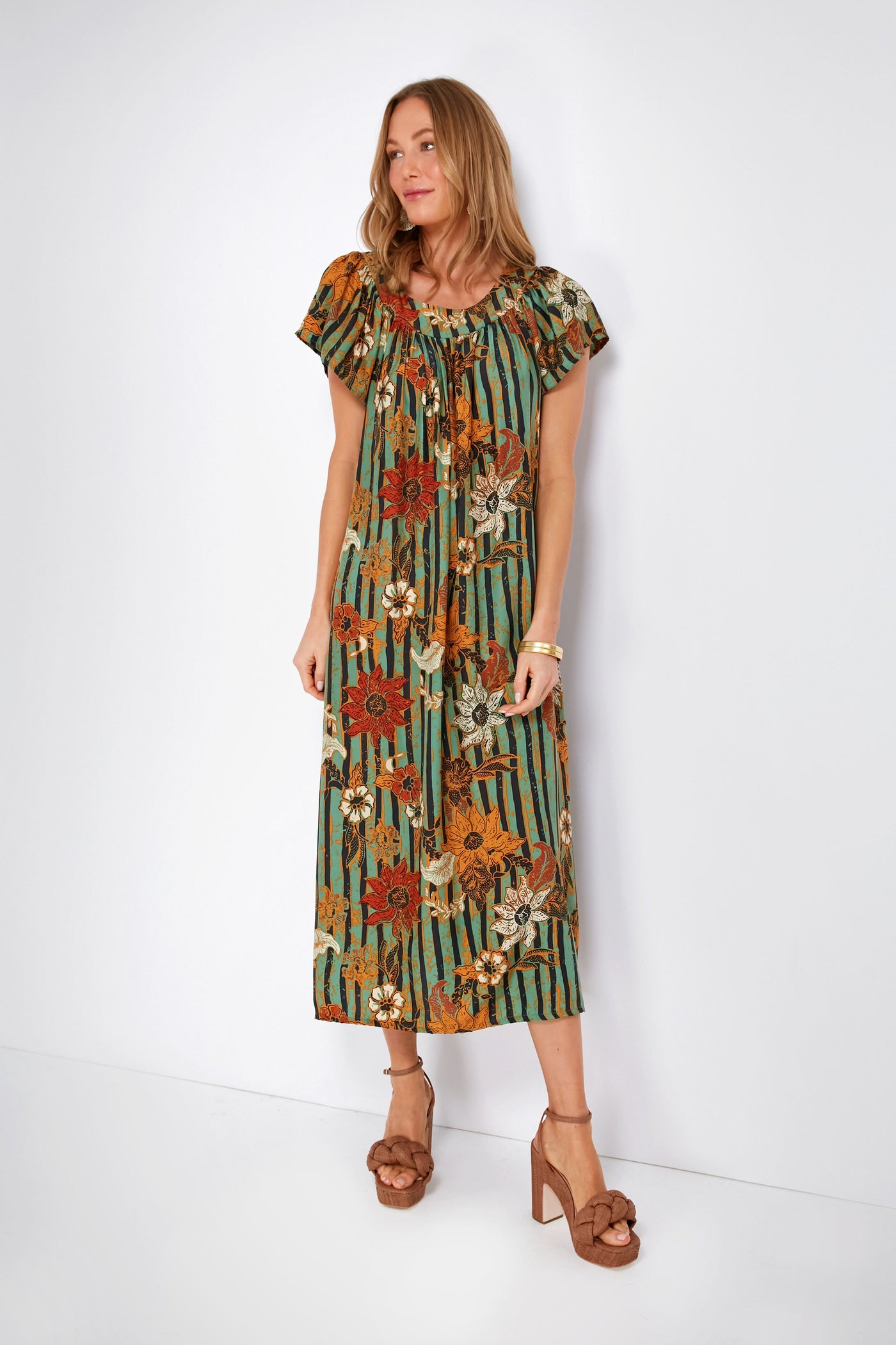 A woman wears a Natalie Martin green and orange floral striped batik-print dress with short sleeves, paired with brown heeled sandals.