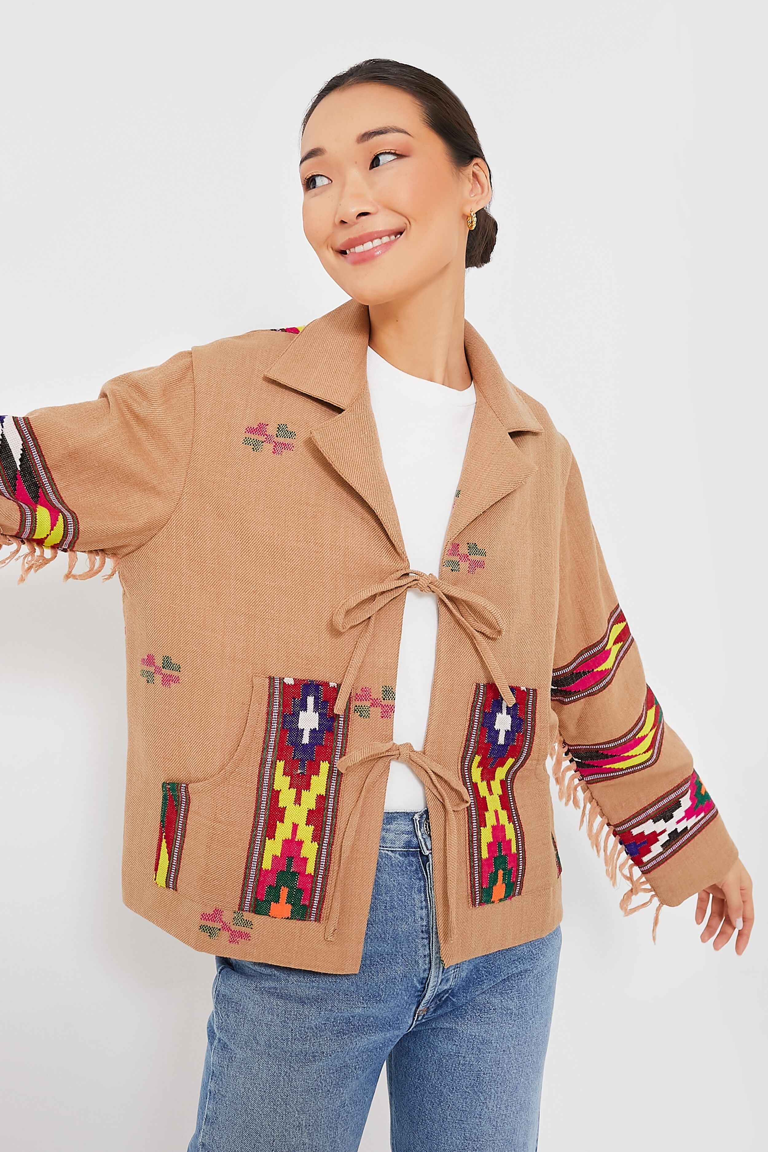 A person wears an Alix of Bohemia tan Western jacket with colorful geometric patterns and handwoven wool details, paired with a white shirt and blue jeans, standing with one arm raised in a boho-chic look.
