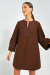 A woman wears Tuckernuck's Brown Tahlia Dress, a long-sleeve A-line mini dress featuring brown fabric with embroidered and eyelet details on the chest and sleeves.