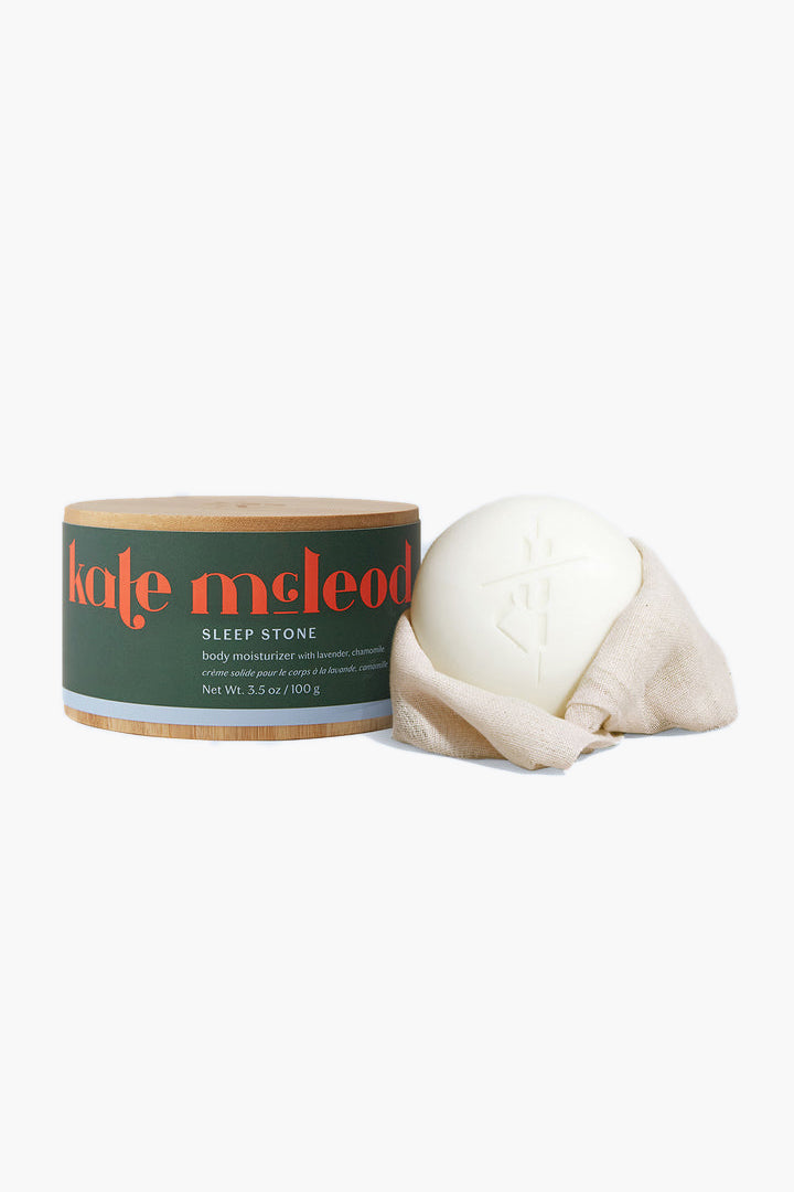 A round Kate McLeod Sleep Stone container sits next to a white, round solid body moisturizer bar, partially wrapped in beige cloth. This waterless lotion is a perfect addition to your nighttime routine.