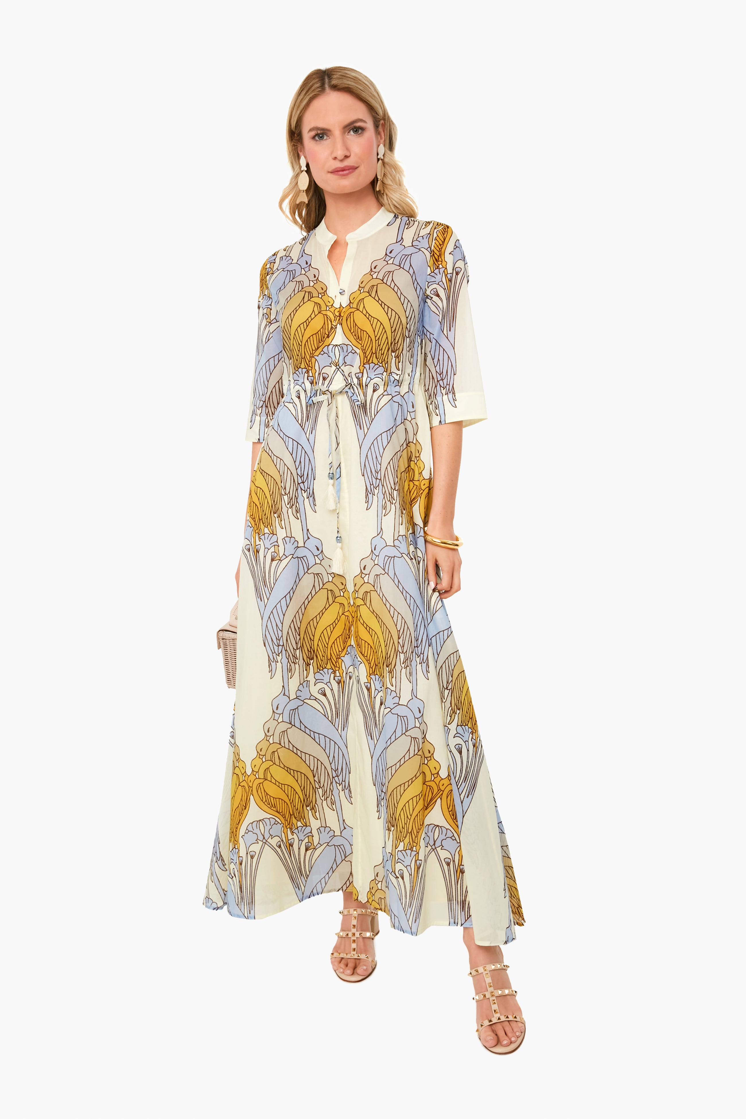 A woman wears a Tory Burch flowy, ankle-length cotton voile dress with a blue and yellow leaf print, paired with sandals and holding a small clutch.