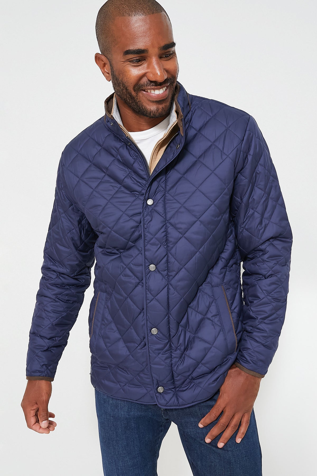 A man in a dark blue Peter Millar travel coat, light shirt, and blue jeans stands smiling and gazing to the side.