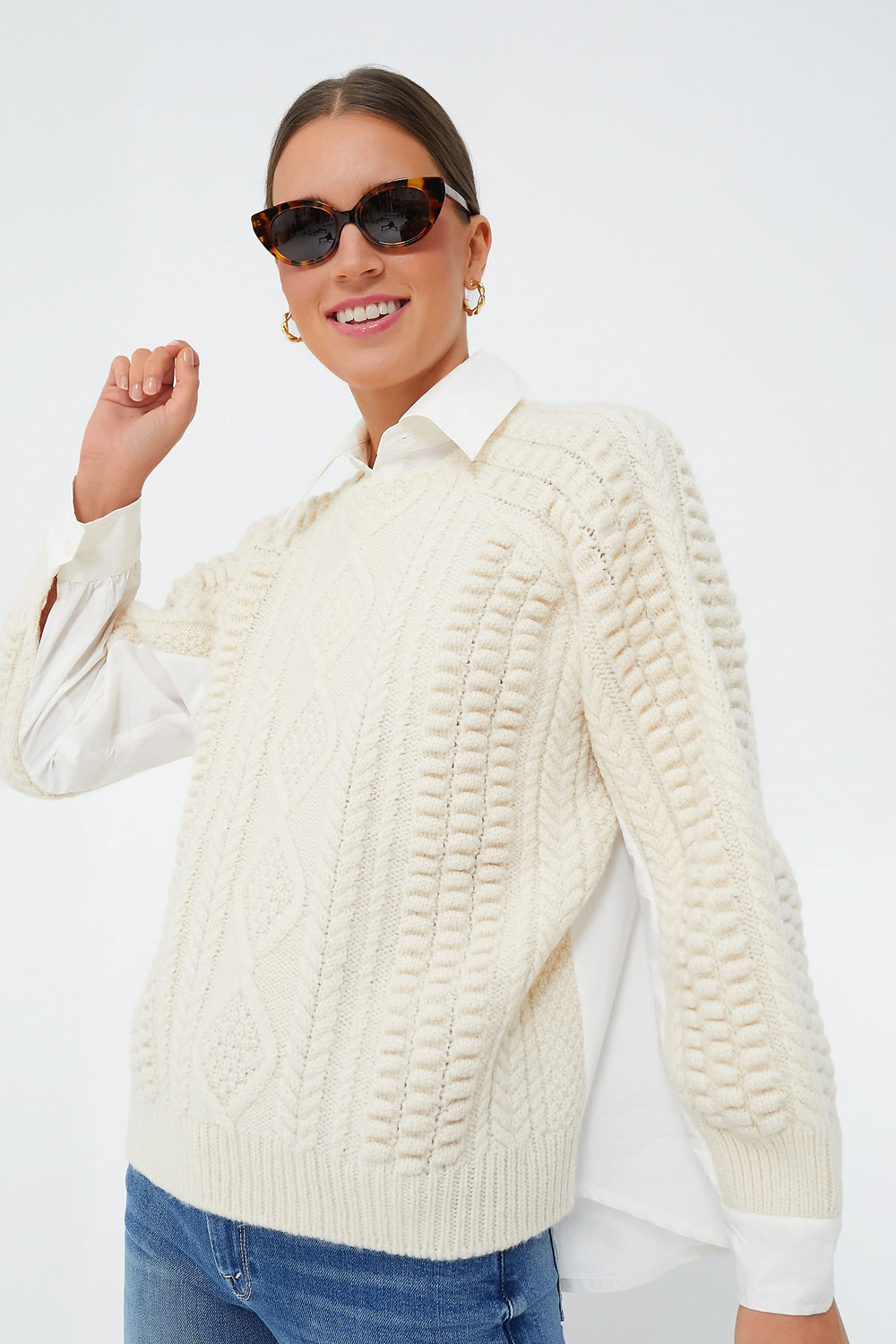 A woman in Sea New York tortoiseshell sunglasses, a mixed media white collared knit cotton shirt layered under a cable long sleeve sweater, and blue jeans poses confidently.
