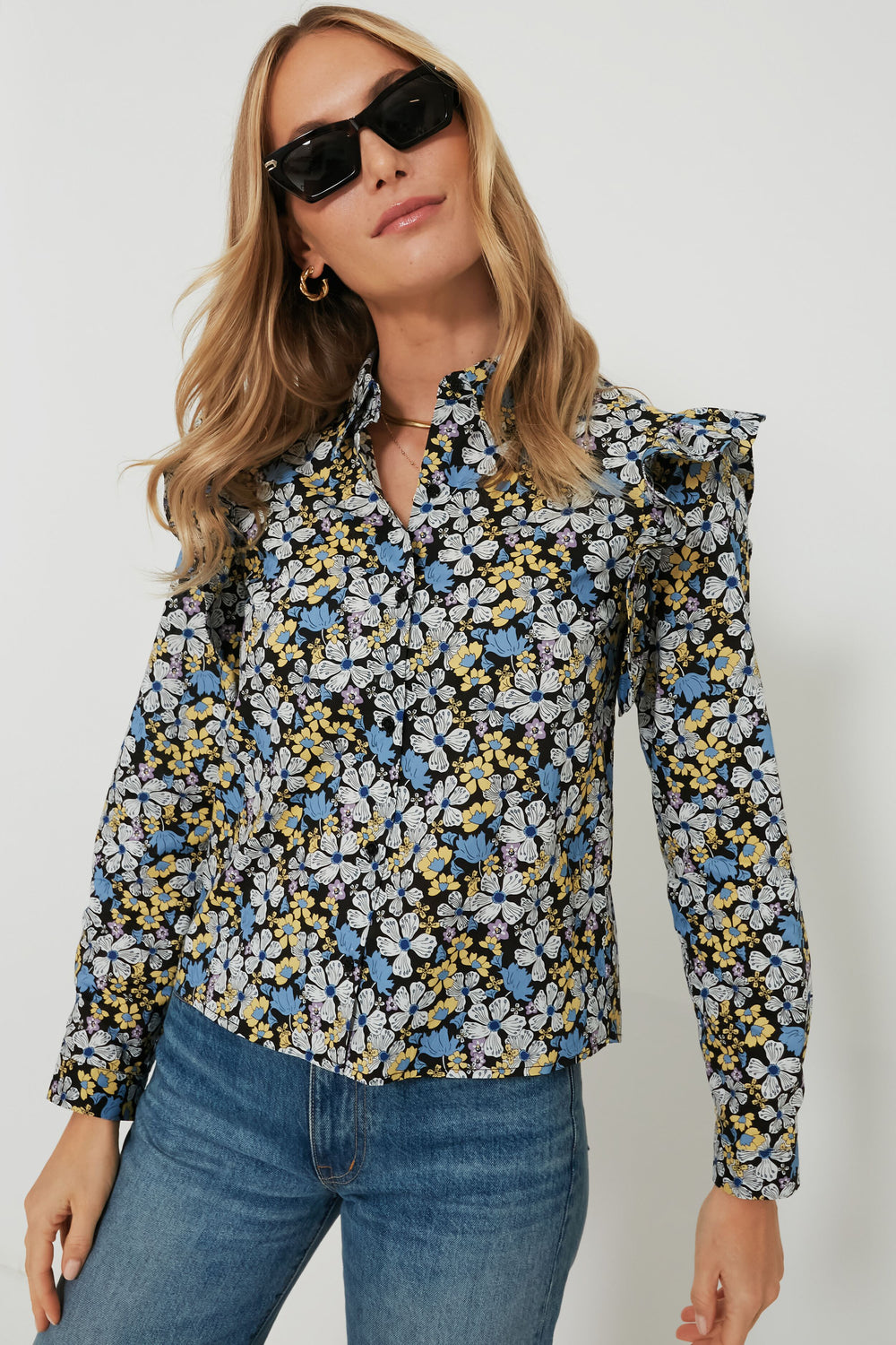 A woman stands confidently, wearing Hyacinth House sunglasses and a long-sleeve floral blouse with ruffle details, paired with blue jeans.