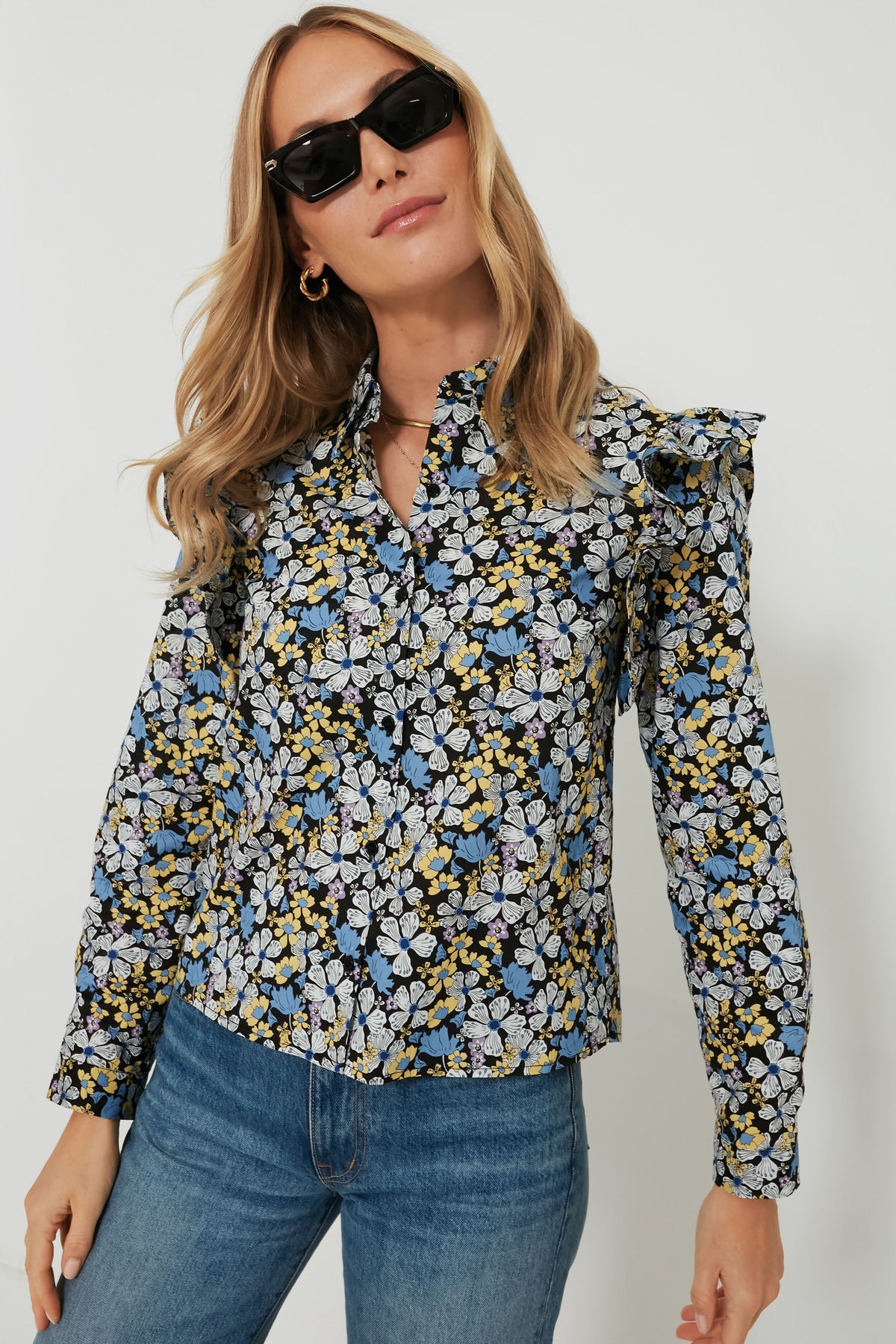A woman stands confidently, wearing Hyacinth House sunglasses and a long-sleeve floral blouse with ruffle details, paired with blue jeans.