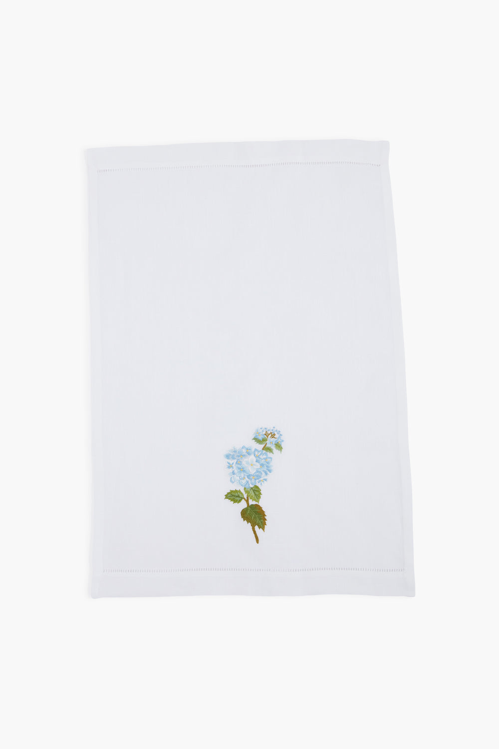A white linen guest towel by Tuckernuck Home, featuring a hand-embroidered blue flower with green leaves in one corner.