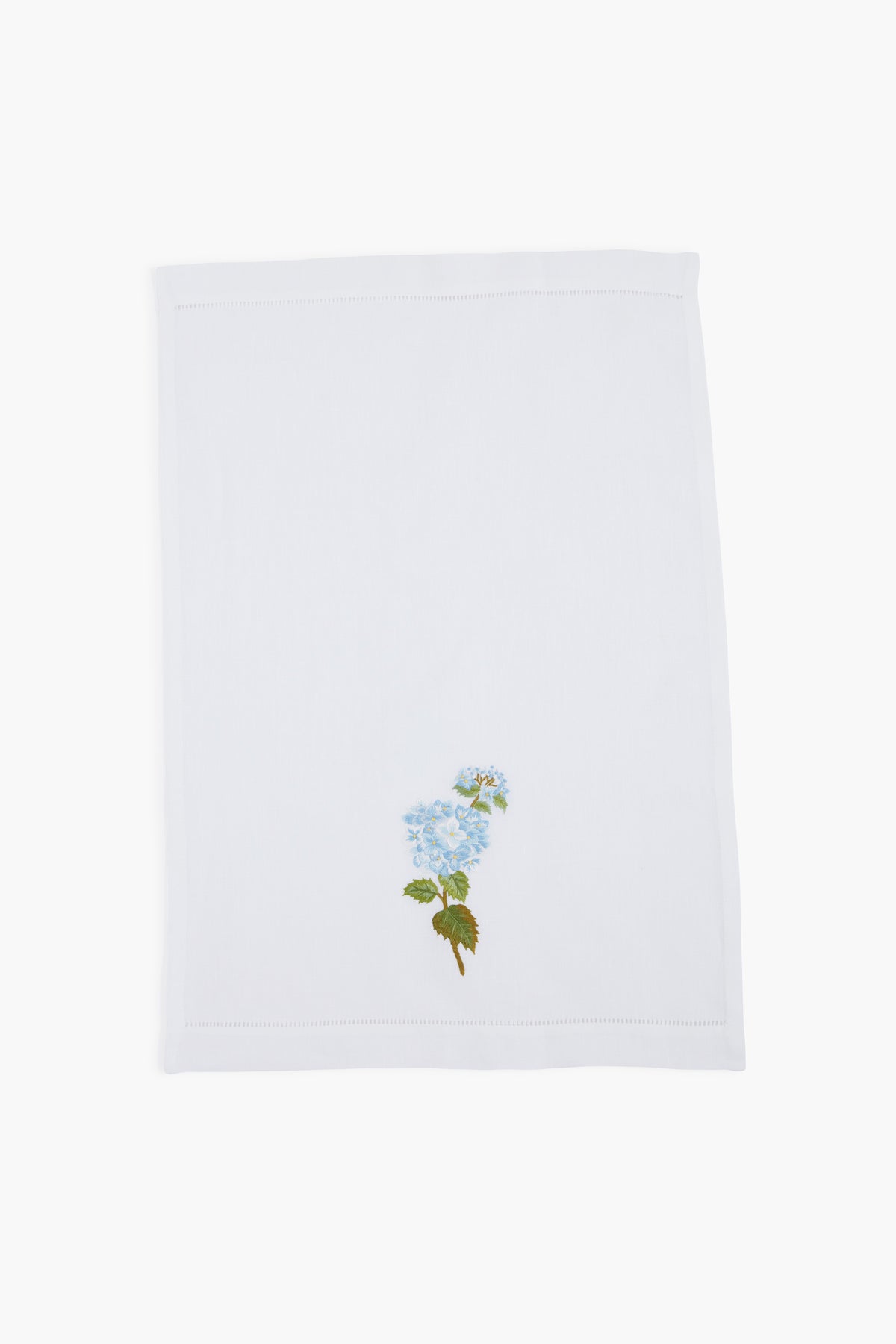 A white linen guest towel by Tuckernuck Home, featuring a hand-embroidered blue flower with green leaves in one corner.