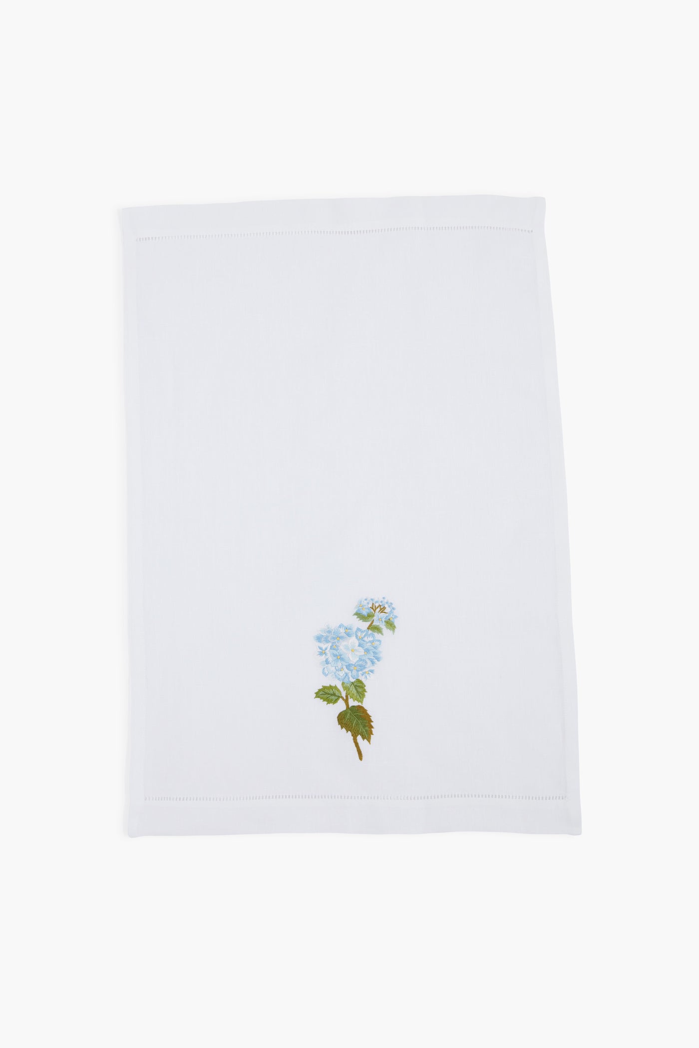 A white linen guest towel by Tuckernuck Home, featuring a hand-embroidered blue flower with green leaves in one corner.