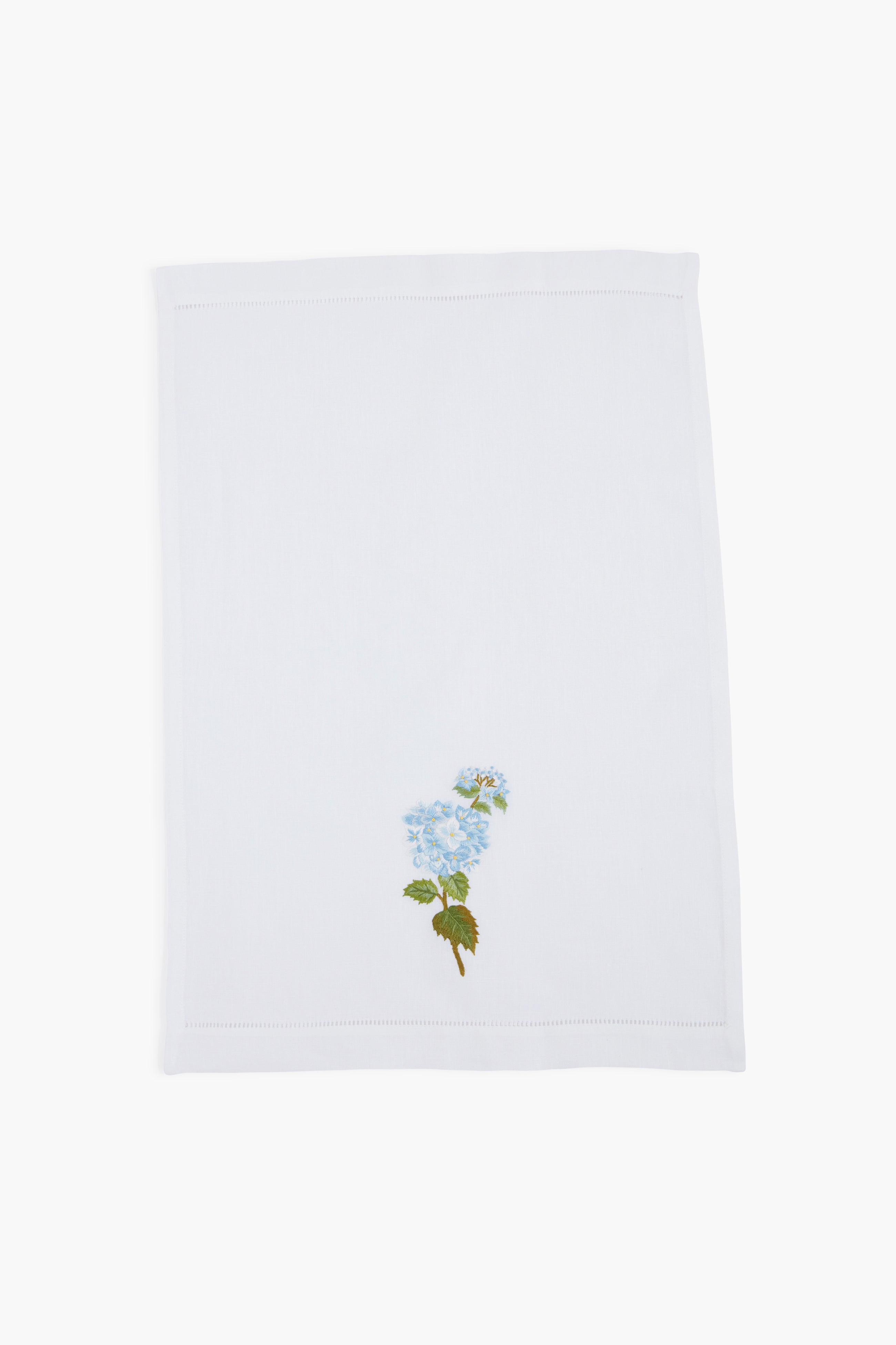 A white linen guest towel by Tuckernuck Home, featuring a hand-embroidered blue flower with green leaves in one corner.