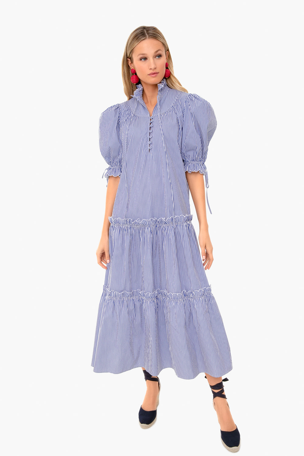 Woman in a Hunter Bell Blue Stripe Holloway Dress with puffed sleeves and ruffle accents, styled with black espadrille shoes and bold red earrings for an effortlessly timeless coastal look.