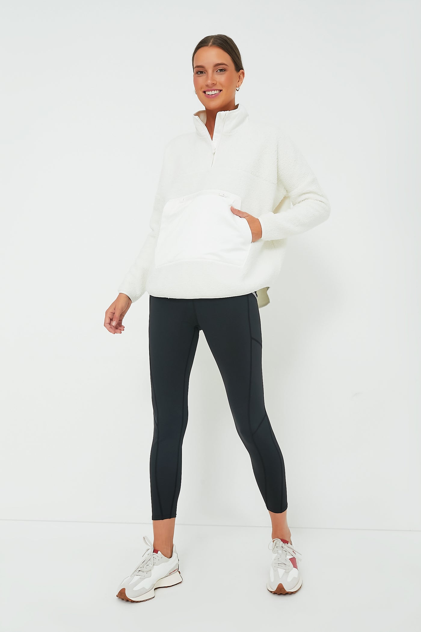 A woman stands against a white background wearing a Tnuck Sport White Fleece Cady Quarter Zip, black leggings, and white sneakers, with her hair pulled back and hands by her sides.