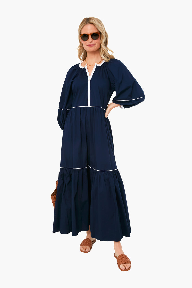 A woman wears a STAUD navy blue maxi dress with white trim and a tiered skirt, brown sandals, sunglasses, and carries a woven bag.