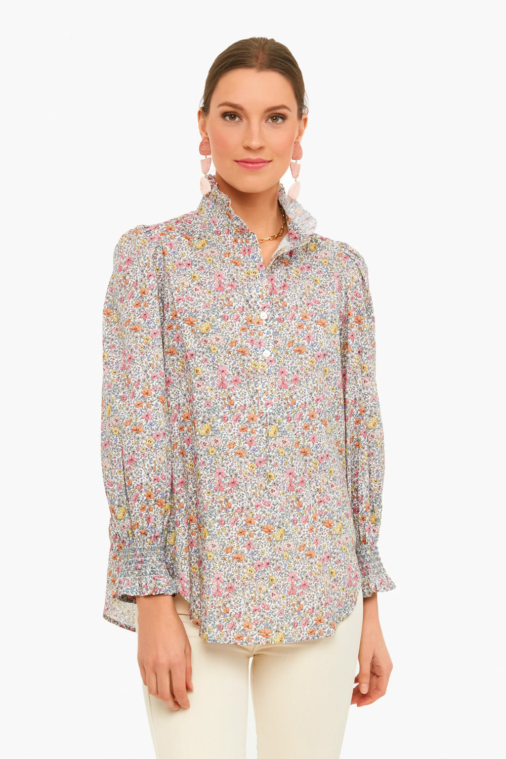 A woman wears the Tuckernuck Teagan Popover Shirt, a floral cotton voile long-sleeve top with ruffle collar, styled with white pants and pink drop earrings.