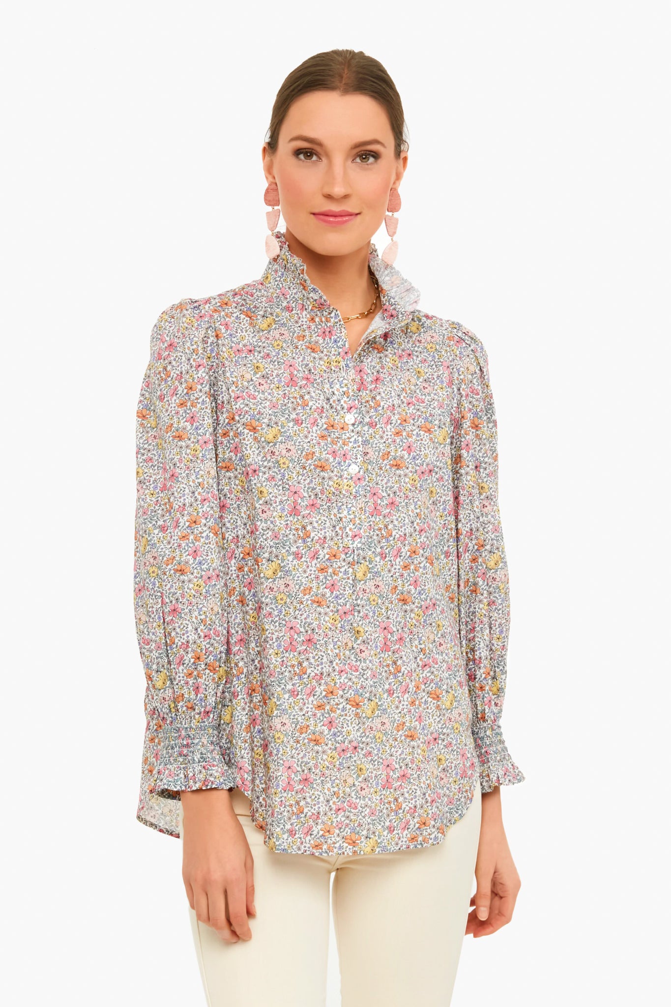 A woman wears the Tuckernuck Teagan Popover Shirt, a floral cotton voile long-sleeve top with ruffle collar, styled with white pants and pink drop earrings.