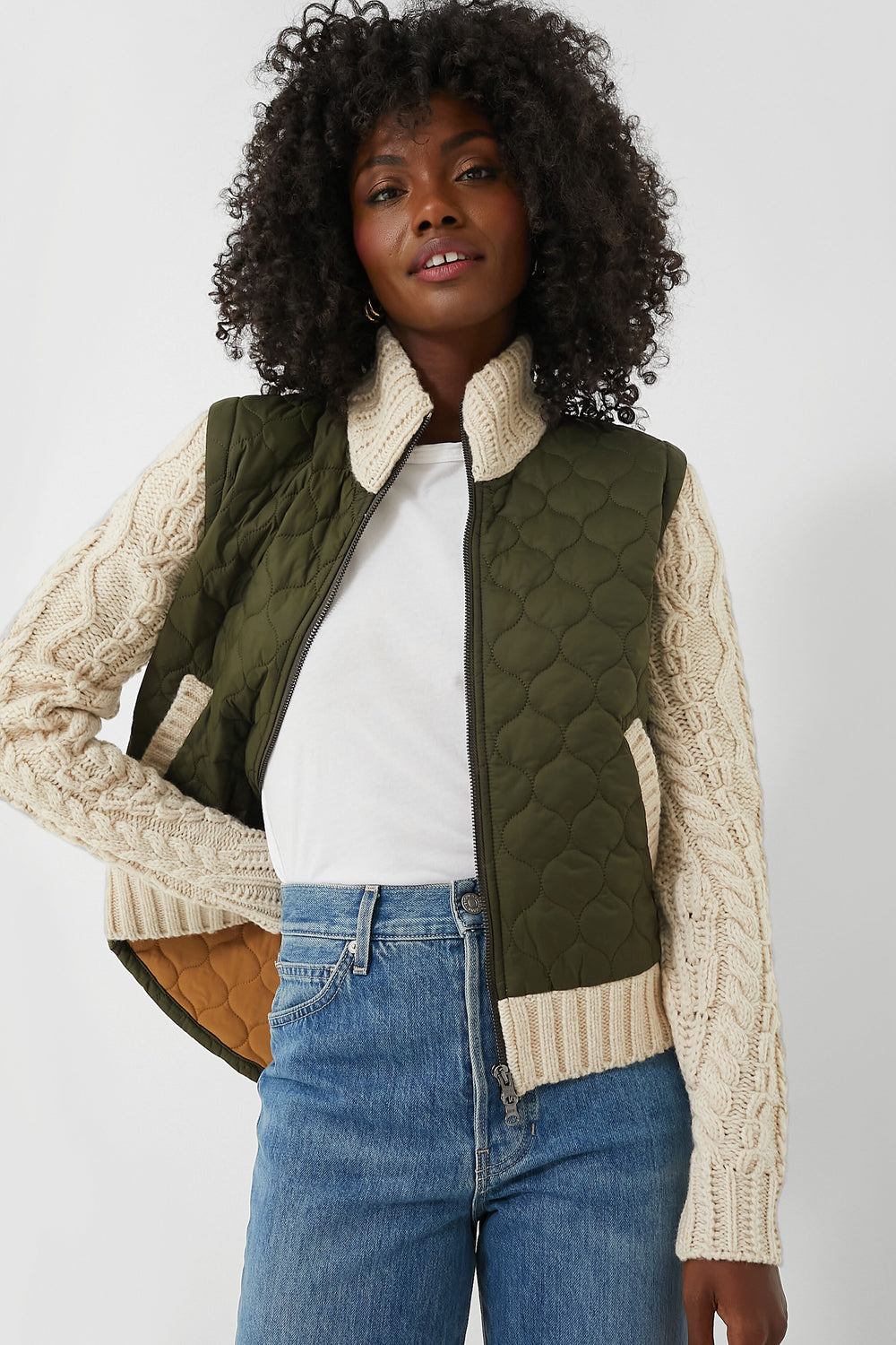A woman wears a white t-shirt, blue jeans, and a Veronica Beard green mixed media jacket with cream knit sleeves, inspired by modern bomber styles.