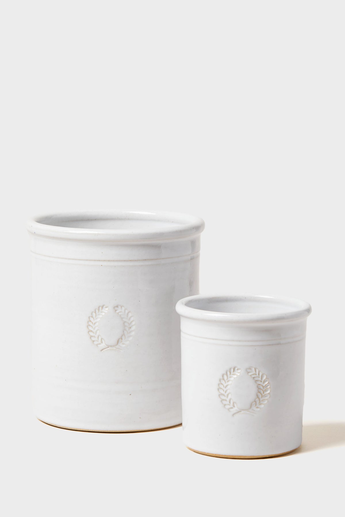 Two white ceramic canisters of different sizes by Farmhouse Pottery, Inc, feature embossed wreath designs on the front and are arranged side by side.