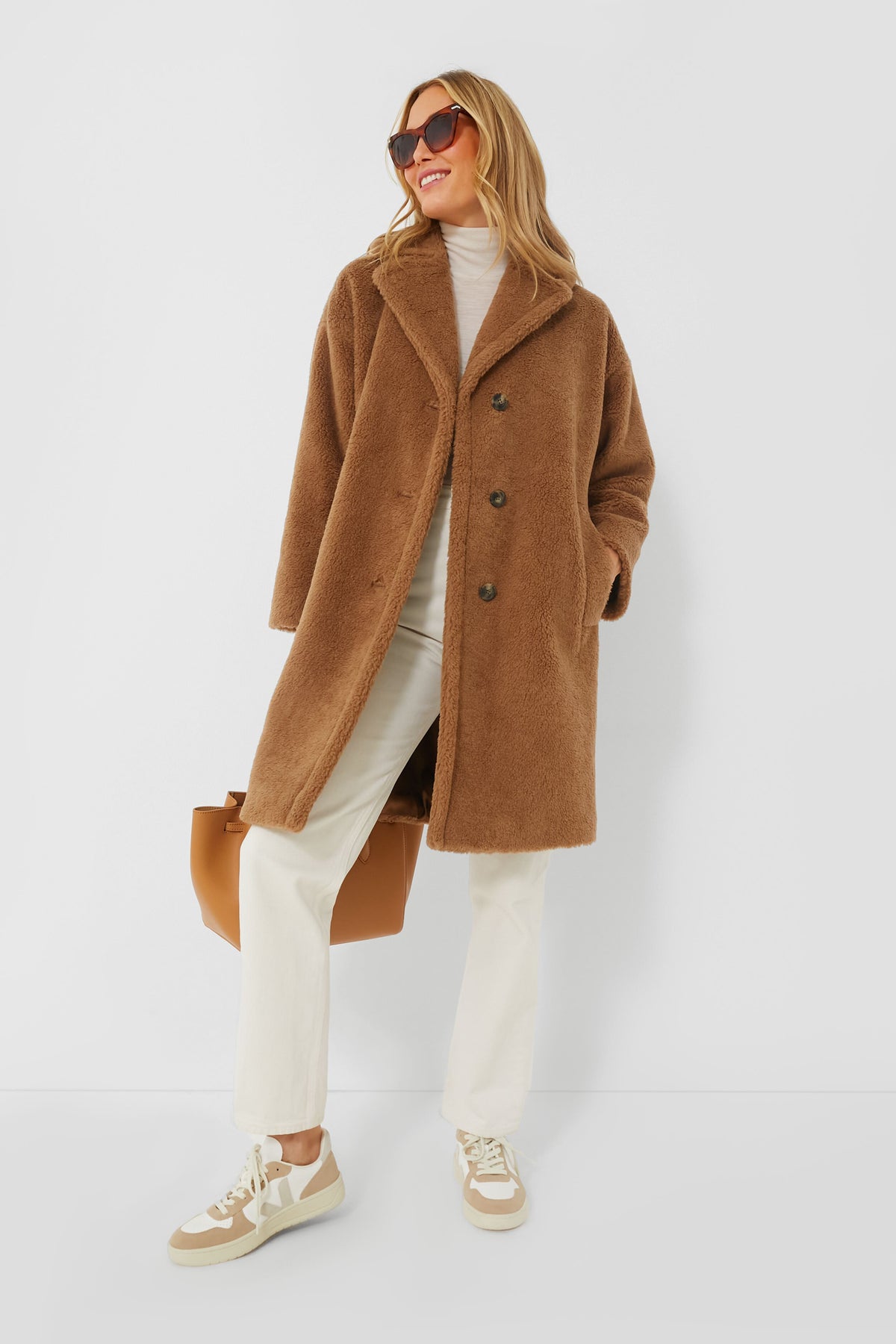 A woman in a camel Weekend Max Mara Veber coat, cream turtleneck, light pants, beige sneakers, and sunglasses stands holding a tan bag.