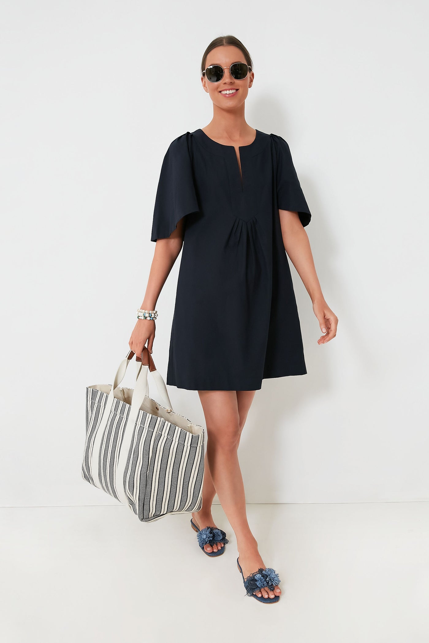 A person in a Pomander Place black mini dress with flutter sleeves, sunglasses, and slide sandals carries a large striped tote bag.