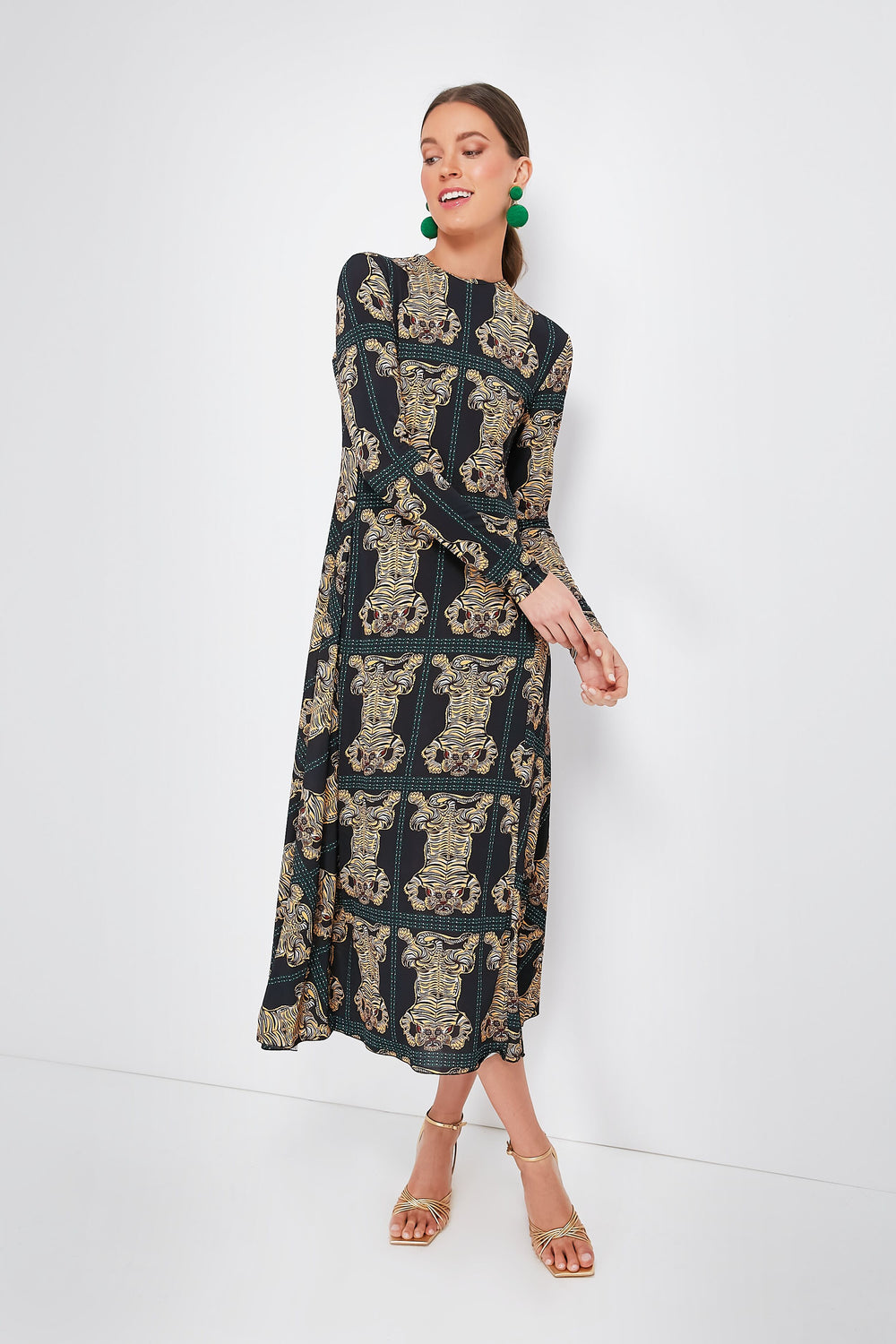 A woman wears a Tiger Tiles Black Drifter Dress by La DoubleJ, featuring long sleeves, an ankle-length gold ornate pattern, and gold sandals. She looks to the side and smiles.