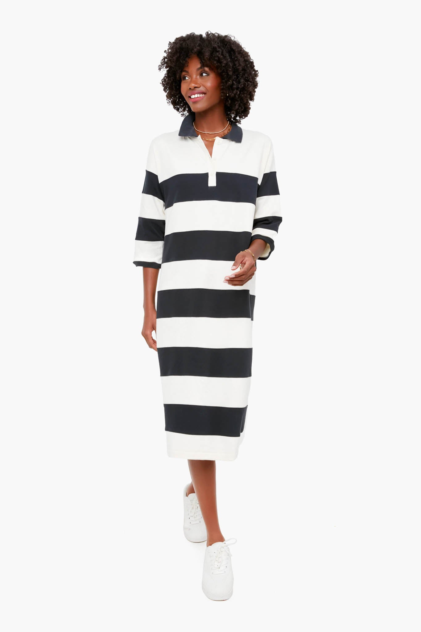 A woman wears THE GREAT.’s black and white striped Rugby Polo Dress with three-quarter sleeves and white sneakers, showcasing a timeless wardrobe staple.