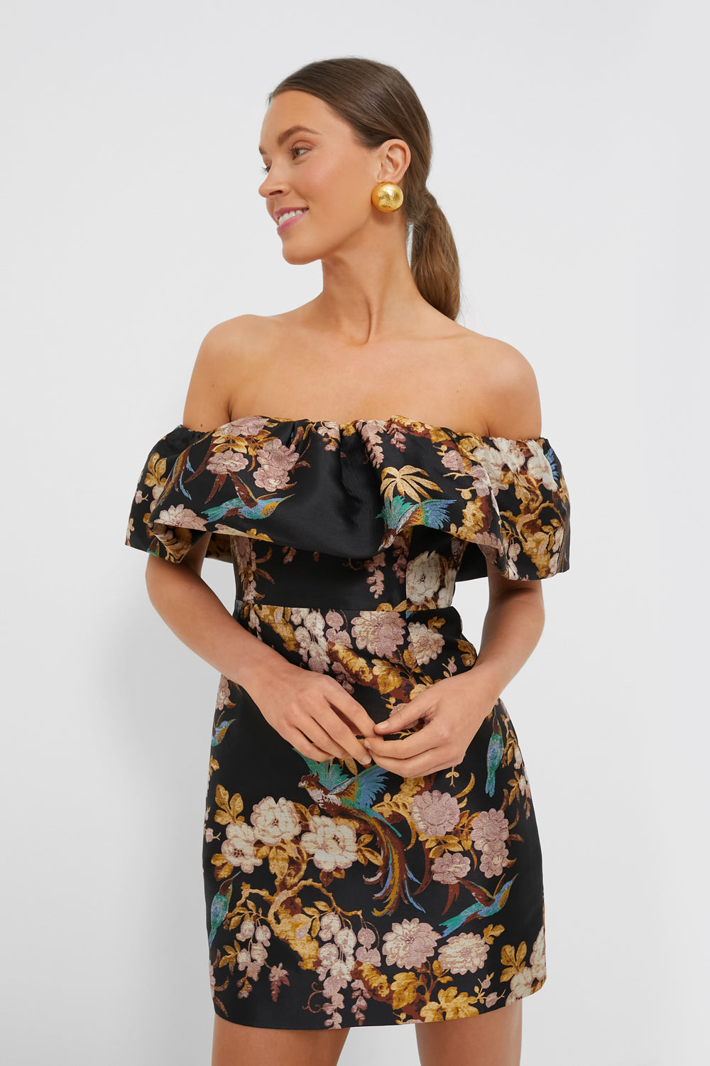 Woman wearing an off-the-shoulder, floral-patterned Black Birds of Paradise Vera Dress by Cara Cara with large gold earrings—perfect for evening occasions.
