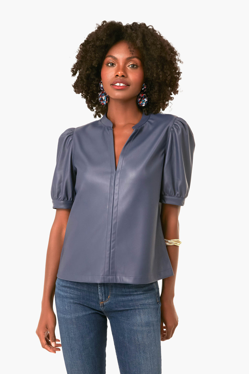 A woman exudes stylish confidence in a BB Dakota x Steve Madden faux-leather top with puff sleeves, paired with blue jeans and statement earrings.