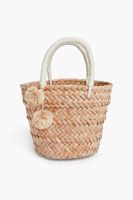 The Kayu woven straw tote bag features two wrapped handles and beige pom-pom accents, expertly crafted with Filipino basket weaving techniques for a timeless, sustainable accessory.