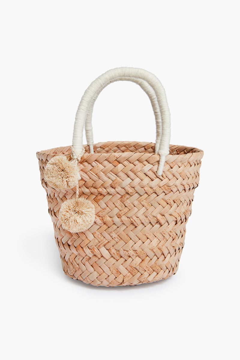 The Kayu woven straw tote bag features two wrapped handles and beige pom-pom accents, expertly crafted with Filipino basket weaving techniques for a timeless, sustainable accessory.