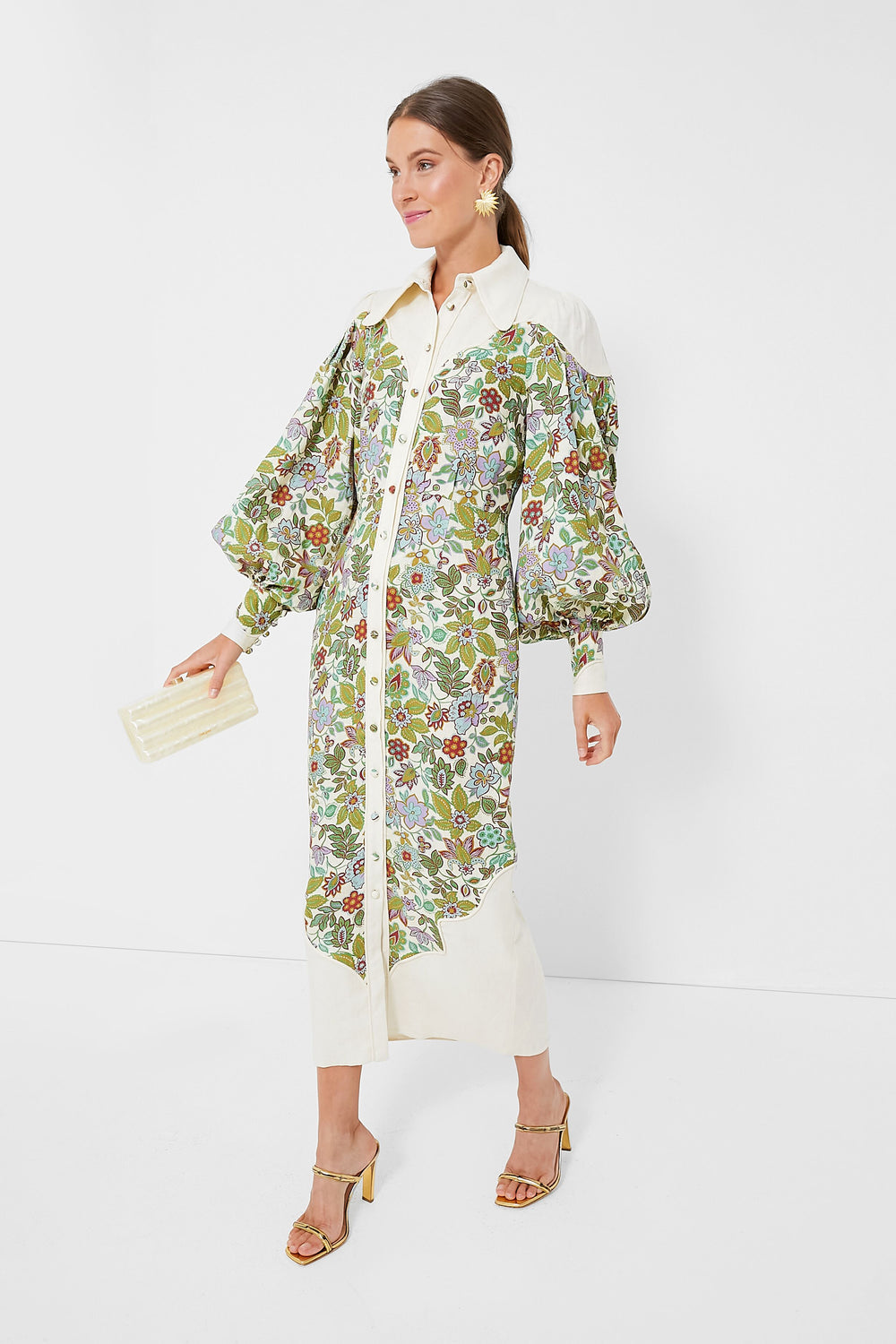 A woman wears an Alemais floral linen midi dress with puffed sleeves, gold sandals, statement earrings, and carries a cream clutch.