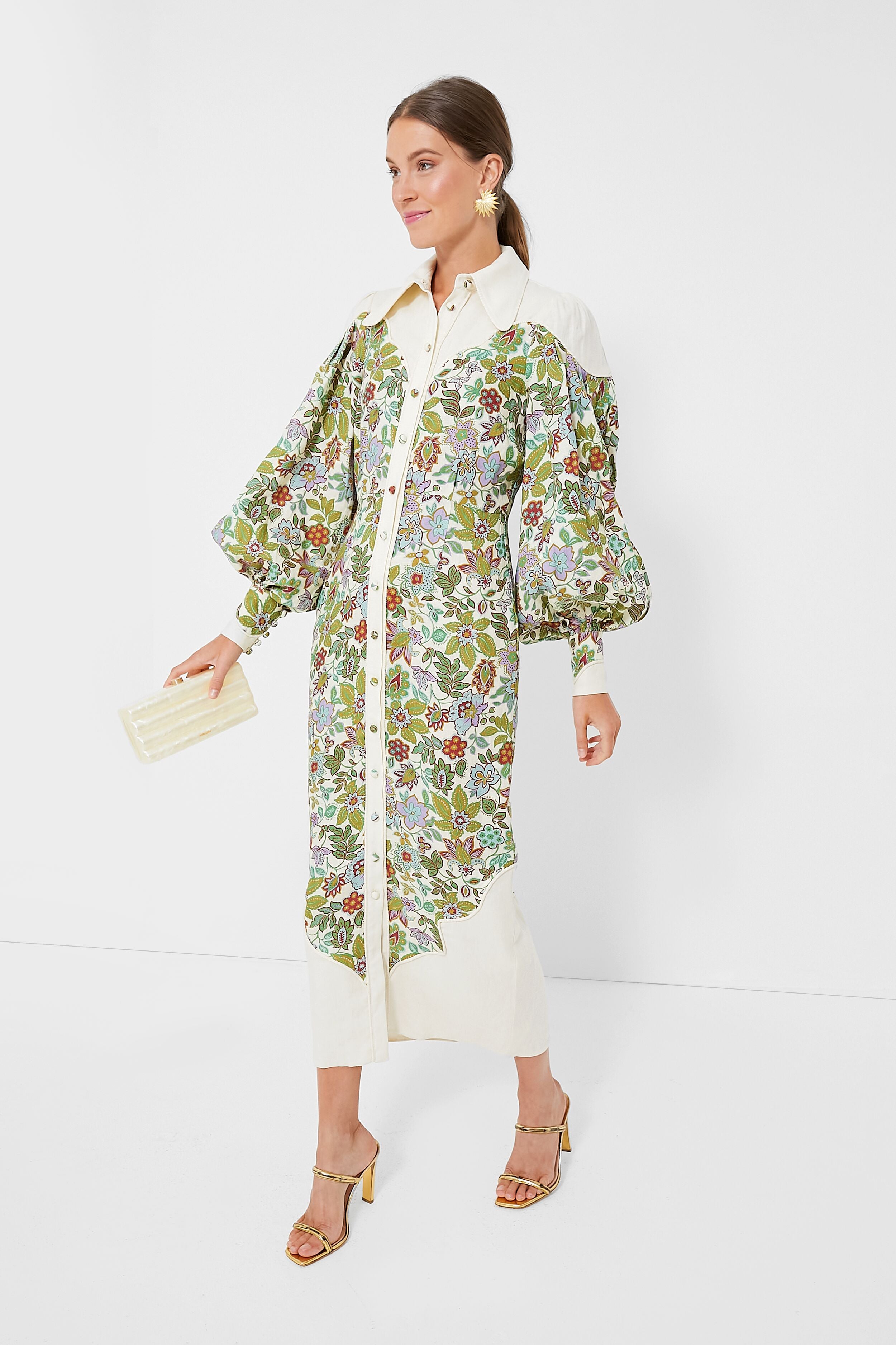 A woman wears an Alemais floral linen midi dress with puffed sleeves, gold sandals, statement earrings, and carries a cream clutch.