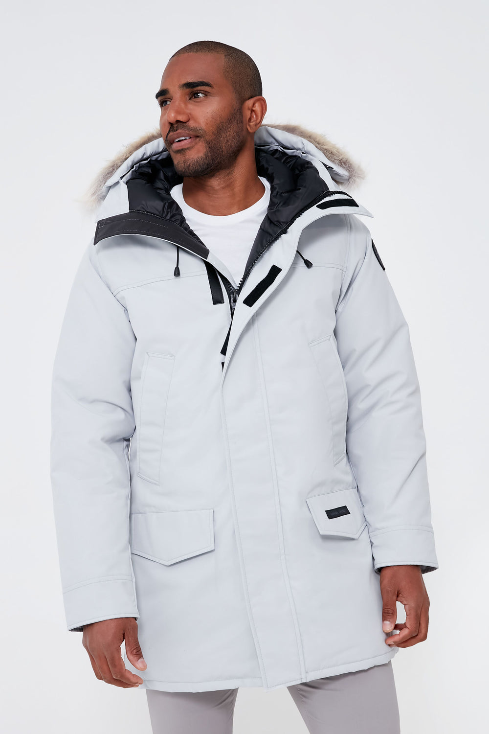 A man in a light gray Canada Goose parka with a fur-trimmed hood stands, glancing to the side.