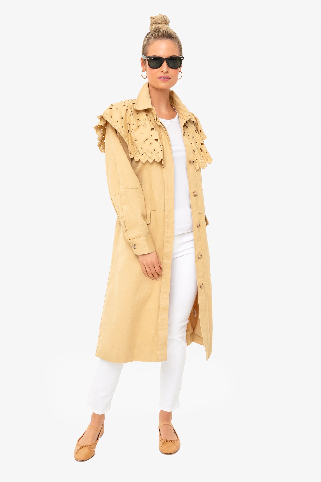 A woman wears a Sea New York beige trench coat with eyelet trim and self-tie belt, styled with a white top and pants, tan flats, sunglasses, and hoop earrings.
