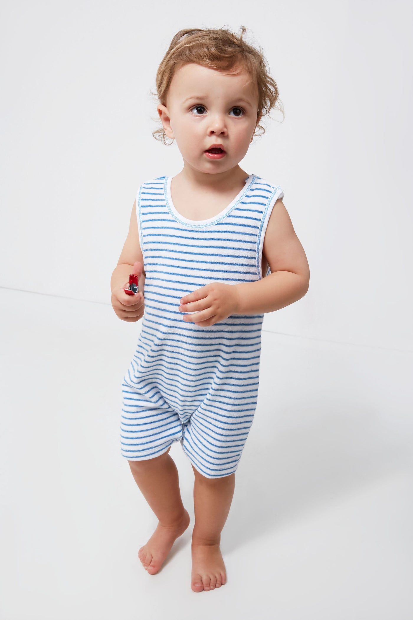 A toddler in a kissy kissy Blue Terry Sleeveless Play Overall stands barefoot on a white floor, holding a small red object.