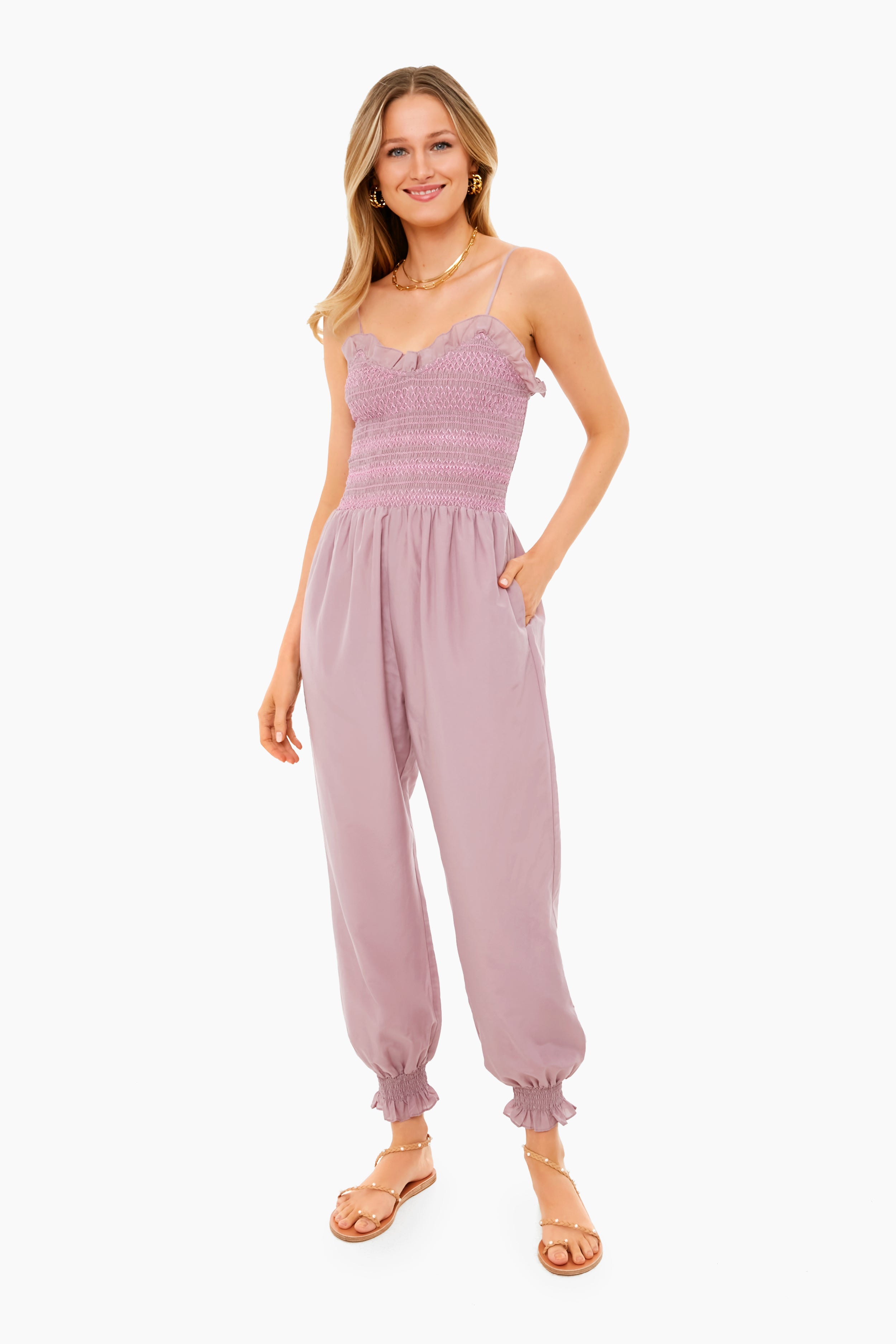A woman smiles with hands in pockets, wearing the Hunter Bell Simone Jumpsuit—a light purple, sleeveless style with a ruffled top, sweetheart neckline, cinched ankles, and crafted from a soft cotton-silk blend for effortless elegance.