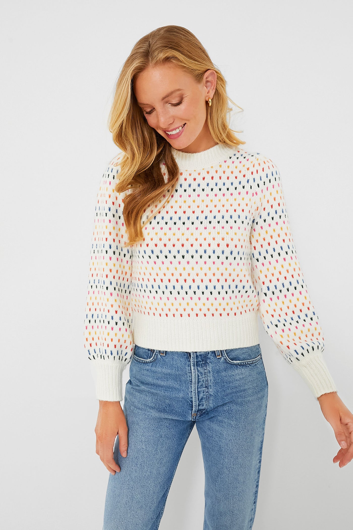 A woman in a Marine Layer white puff sleeve sweater with a multicolored dot pattern and blue jeans looks down with a slight smile.