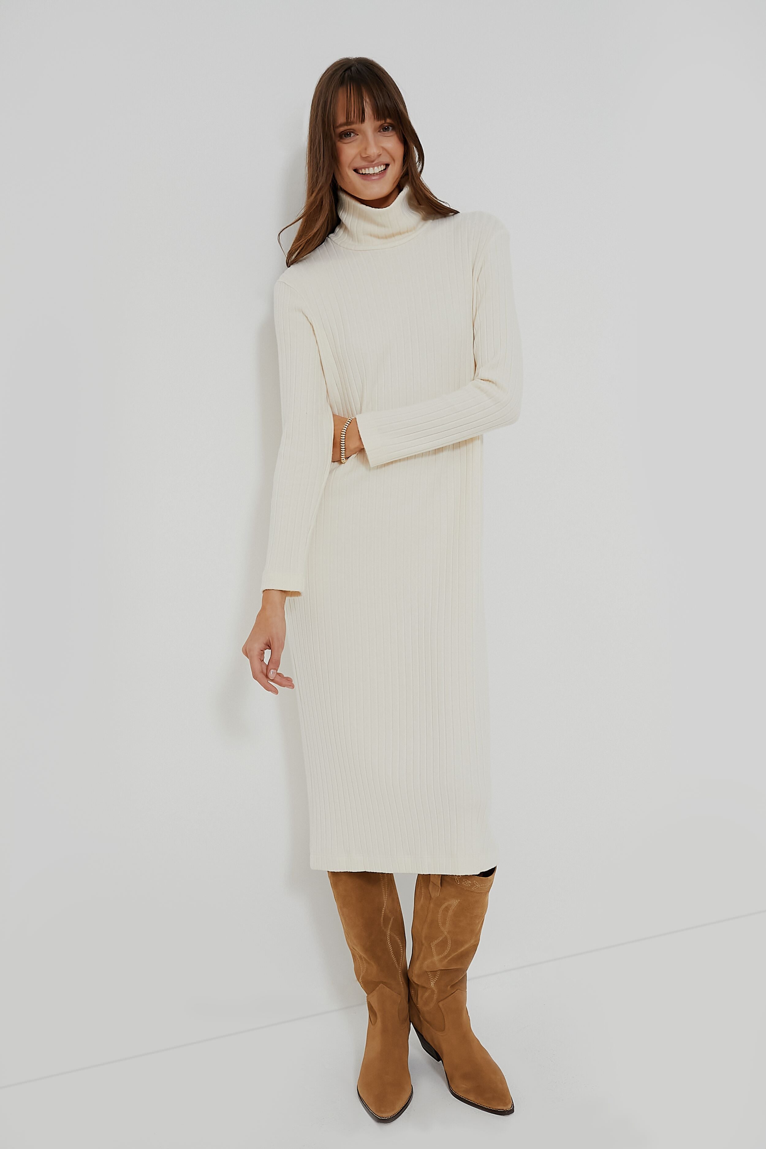 A woman smiles confidently wearing a cream-colored DONNI. organic cotton turtleneck dress with long sleeves and brown knee-high boots.