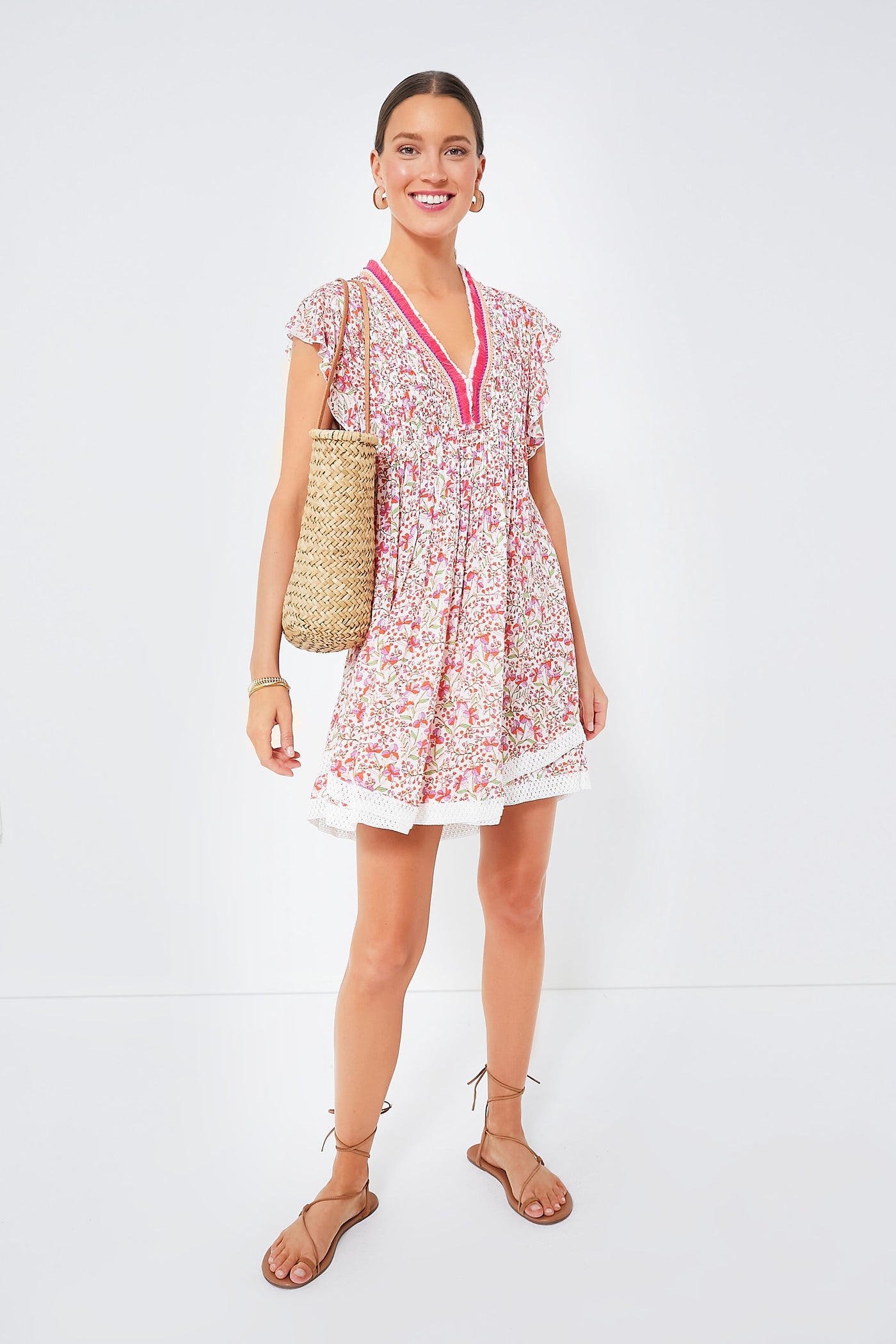 A woman wears a boho-chic Sasha Dress in pink by Poupette St Barth, styled with brown sandals and a woven tote bag.