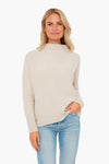 A woman wearing a Vince light beige boiled cashmere sweater and blue jeans stands facing forward with one hand in her pocket.