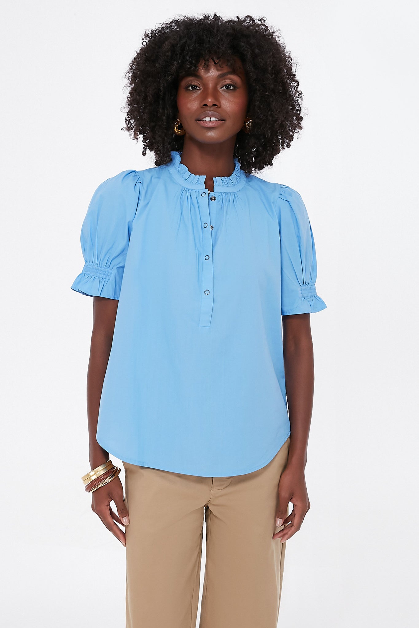 A woman wears an Apiece Apart light blue ruffle collar blouse made from organic cotton, paired with beige pants.