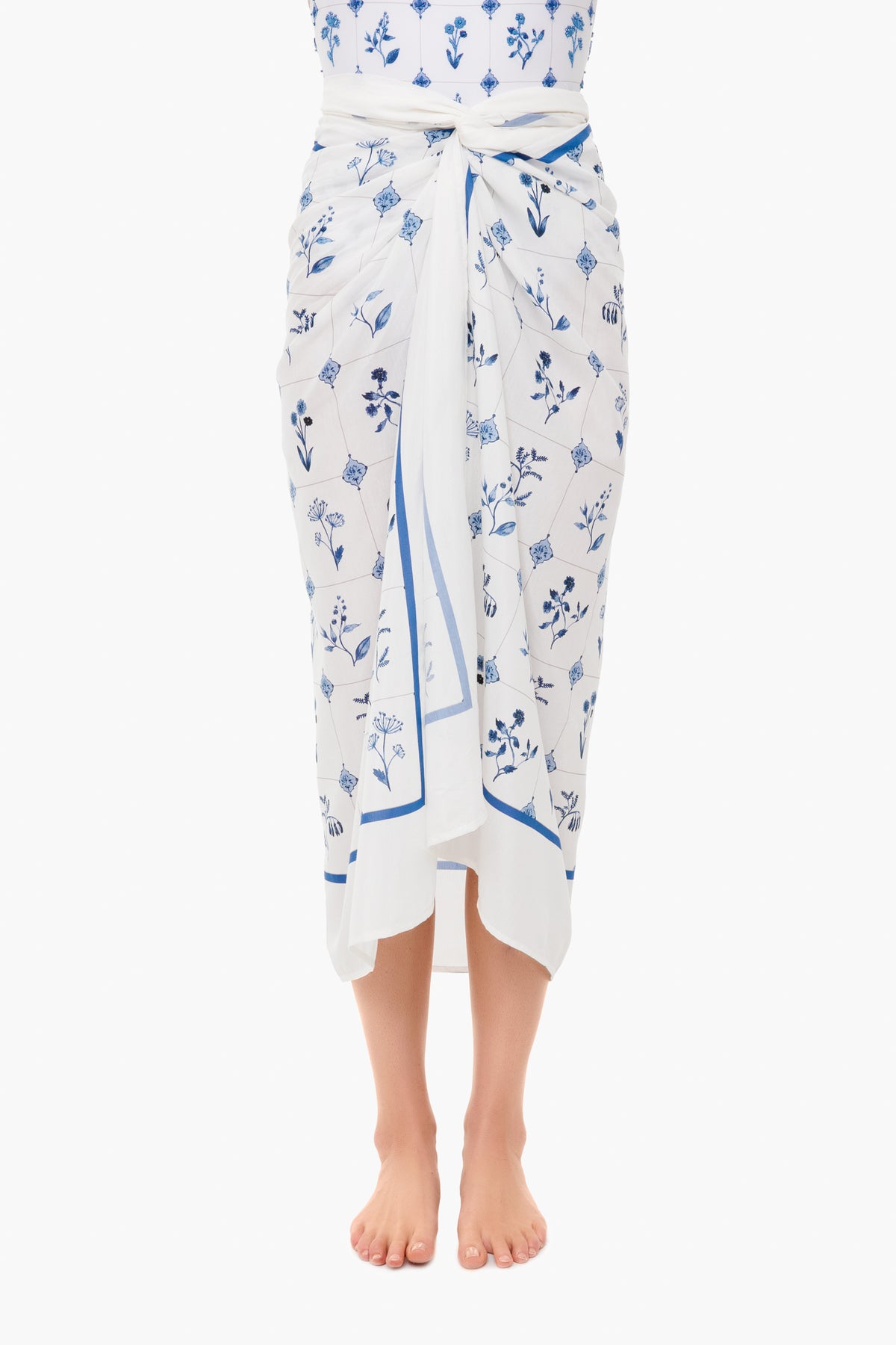 A person stands barefoot, wearing a cotton silk floral pareo by AGUA BY AGUA BENDITA, tied at the waist. The upper body is partially visible.
