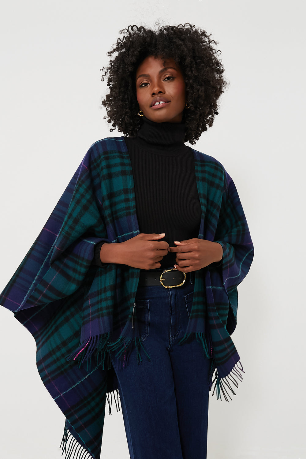 A woman wears a black turtleneck, blue jeans, and a Vineyard Vines green and navy Blackwatch Blanket Wrap with fringed edges draped over her shoulders.