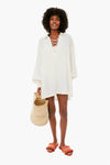 A woman holds a woven basket bag, wearing a WeWoreWhat off-white lace-up caftan as a beach coverup with orange slide sandals.