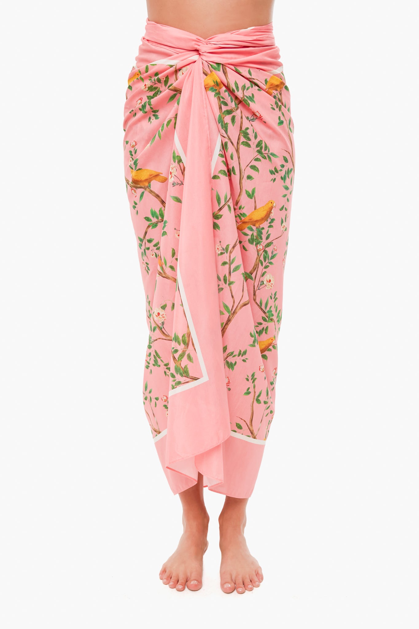 A barefoot person wears a pink floral-patterned cotton-silk pareo by AGUA BY AGUA BENDITA, tied at the waist. Expertly crafted by Colombian artisans, it merges traditional design with modern flair.