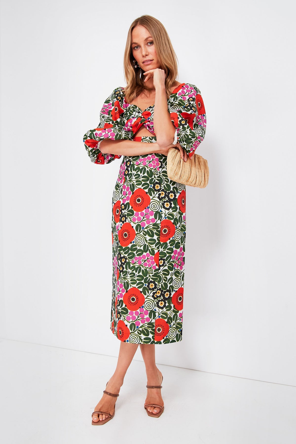 A woman wears the RHODE Aura Blossom Noa Dress, a vibrant floral midi, styled with tan sandals and a woven handbag.