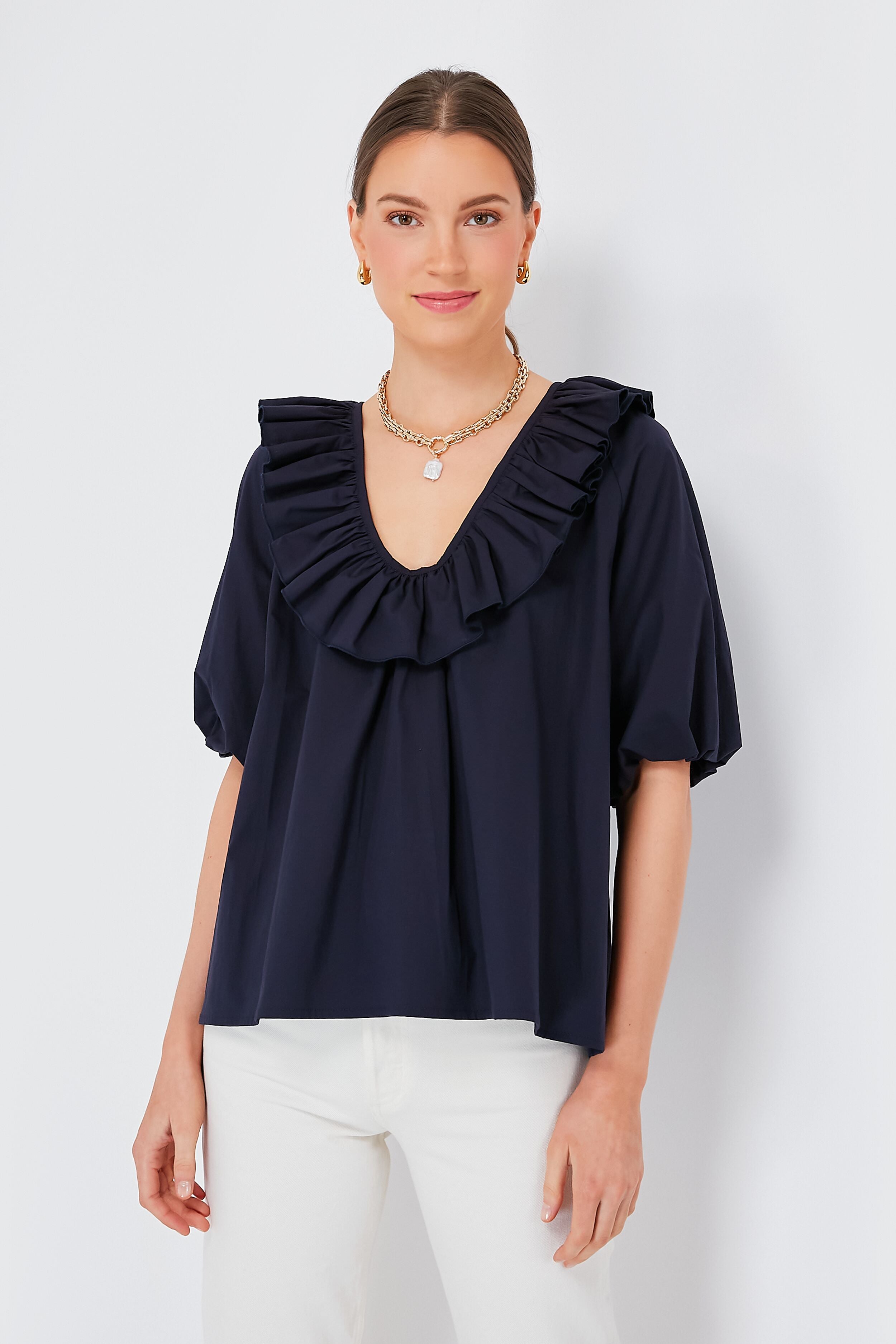 A woman wears a Monica Nera Navy Nancy Top, a ruffled sustainable cotton blouse with short sleeves, paired with white pants.