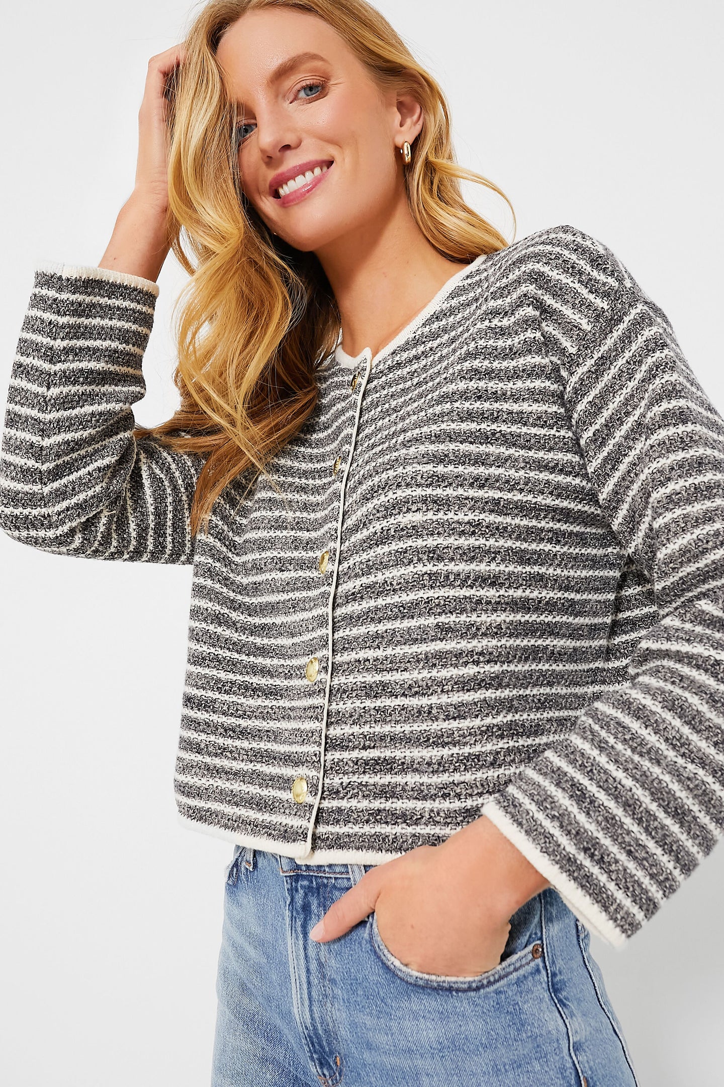 Woman wearing a Barbour black and white striped knitted cardigan with blue jeans, smiling and touching her hair.